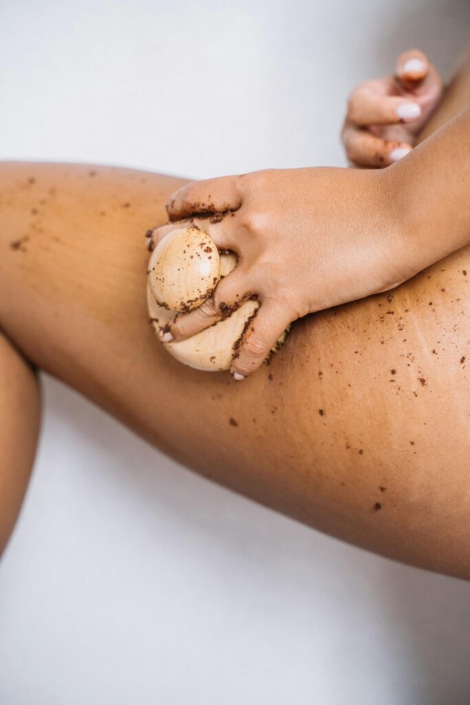 A hand applies coffee scrub on the leg for skin exfoliation in a spa-like setting.