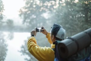 A man in a yellow jacket takes a photo of a misty lake with his smartphone in the morning outdoors.