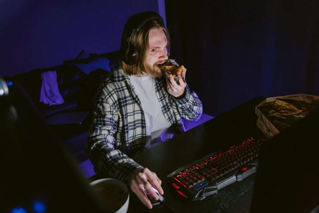 A young man playing video games at night while eating pizza in a dimly lit room.