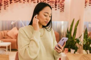 Asian woman enjoying music on wireless earphones while using smartphone indoors.